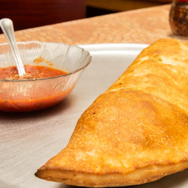 Golden-brown calzone on a metal tray with a clear bowl of marinara sauce and a spoon.
