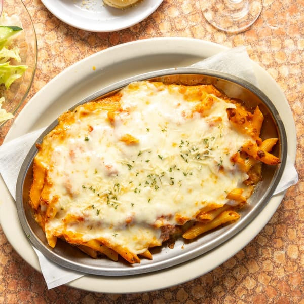 Baked ziti pasta with melted mozzarella cheese and parsley in a metal dish on a patterned table.