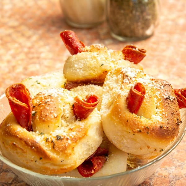 Close-up of golden-brown garlic knots with pepperoni, parmesan cheese, and herbs in a glass bowl.