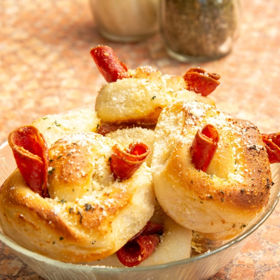 Close-up of golden-brown garlic knots with pepperoni, parmesan cheese, and herbs in a glass bowl.