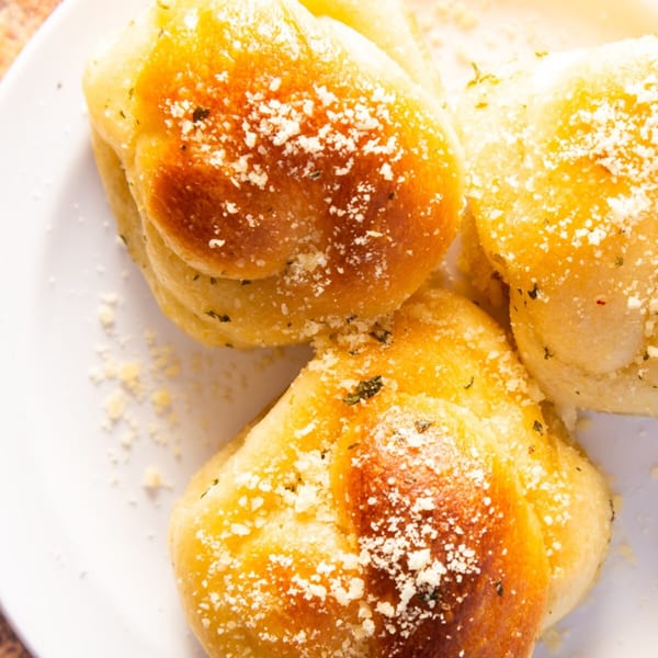 Three golden-brown garlic knots, sprinkled with parmesan cheese and herbs, on a white plate.